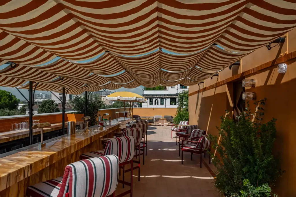 Bar extérieur d’Experimental Roma sous un grand store rayé, avec des tabourets et les toits de Rome
