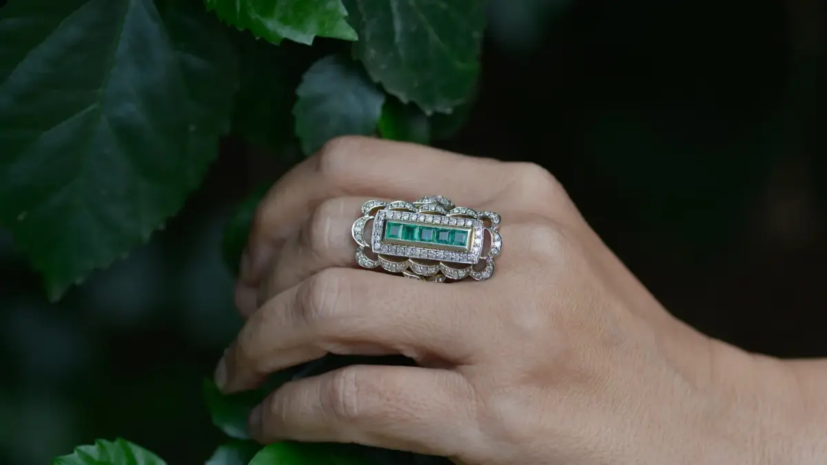 Main portant une bague de fiançailles émeraude et diamants parmi des feuilles vertes