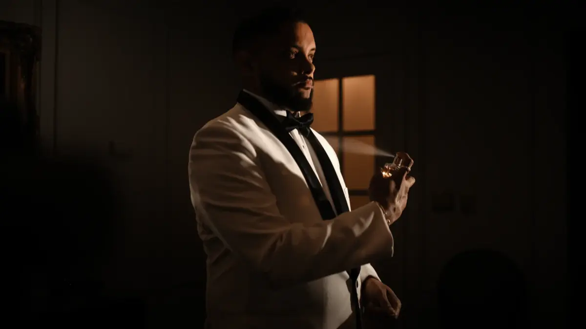 Homme en smoking blanc tenant un flacon de parfum dans une pièce sombre éclairée à la lampe