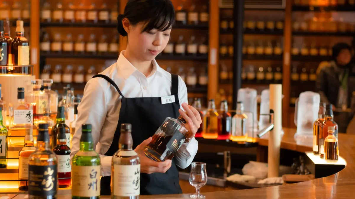 Serveuse présentant une bouteille de whisky japonais derrière un bar garni de bouteilles lumineuses