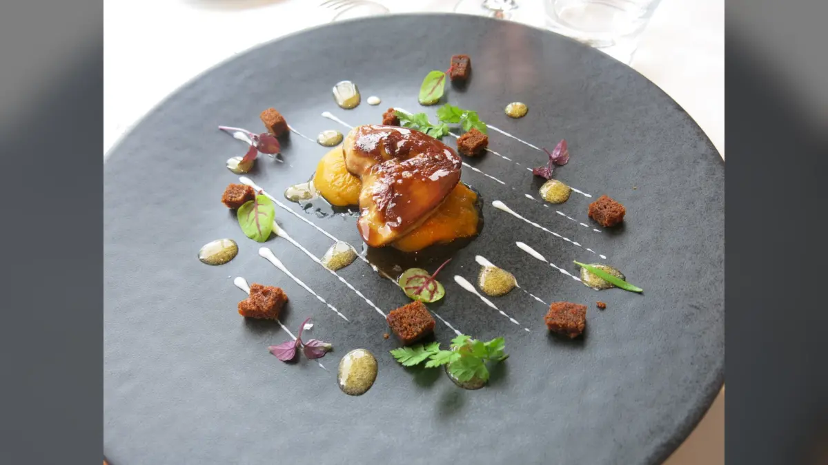 Tranche de foie gras poêlée laquée sur purée, croûtons et herbes dans une assiette noire