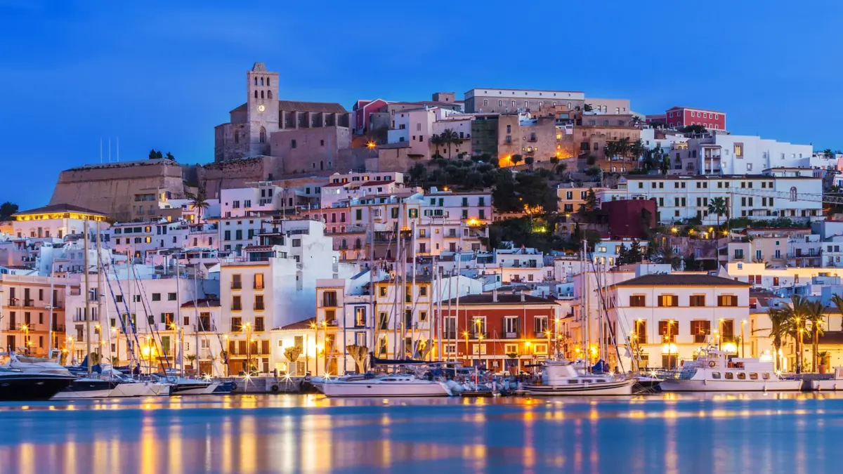 Le port d’Ibiza et la vieille ville de Dalt Vila illuminés à l’heure bleue, voiliers à quai et reflets sur l’eau