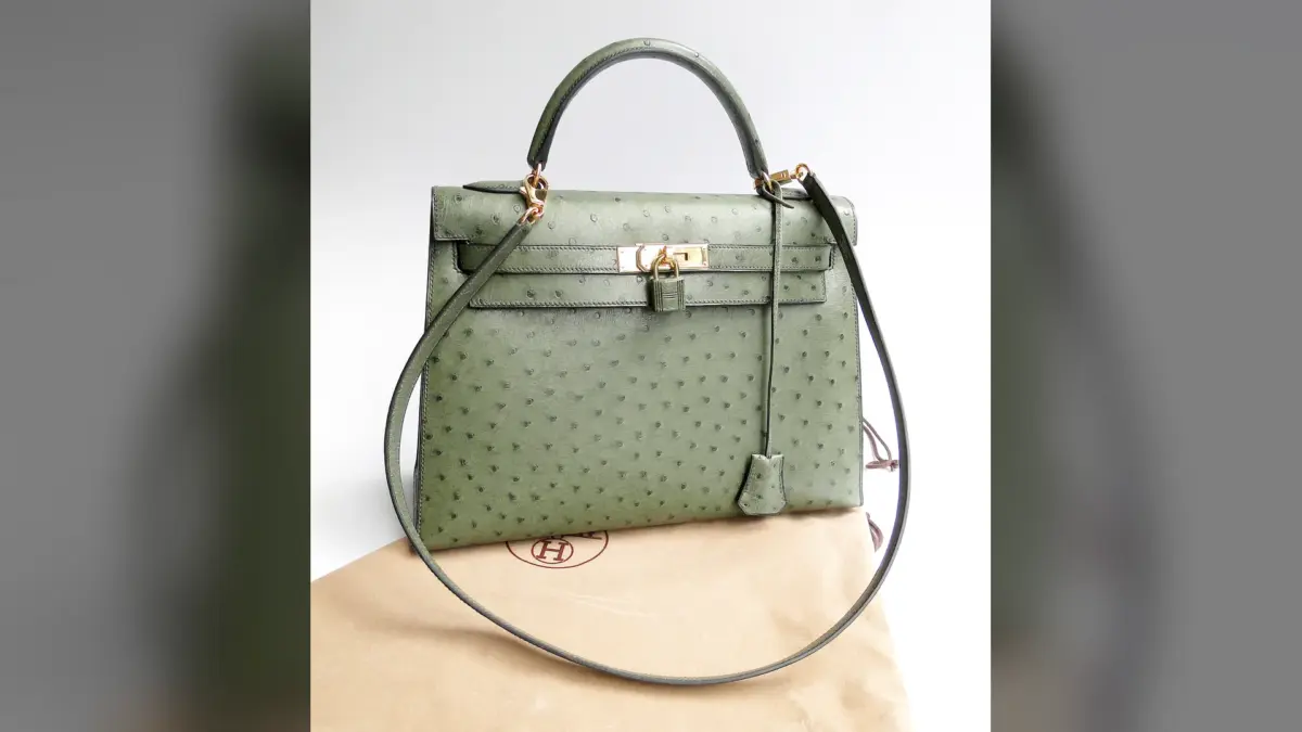 Sac Kelly Hermès vert en cuir d’autruche avec bandoulière, posé sur une housse beige