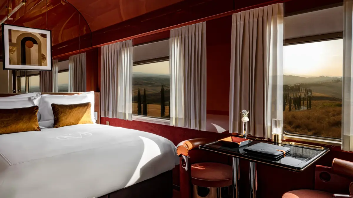 Suite de La Dolce Vita Orient Express aux boiseries laquées rouges, grand lit et fenêtres ouvertes sur les collines de Toscane et leurs cyprès au coucher du soleil